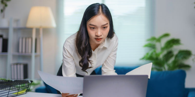 Woman in a white shirt checking some paper infront of her laptop.